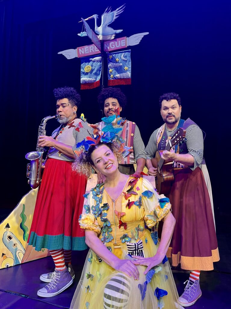 A imagem mostra uma cena vibrante e teatral com quatro artistas em um palco colorido. No centro, uma mulher usa um vestido amarelo adornado com dezenas de borboletas multicoloridas e um elaborado adorno de cabeça com borboletas grandes, criando um visual mágico e fantasioso. Ela parece estar interpretando ou cantando, com gestos expressivos.