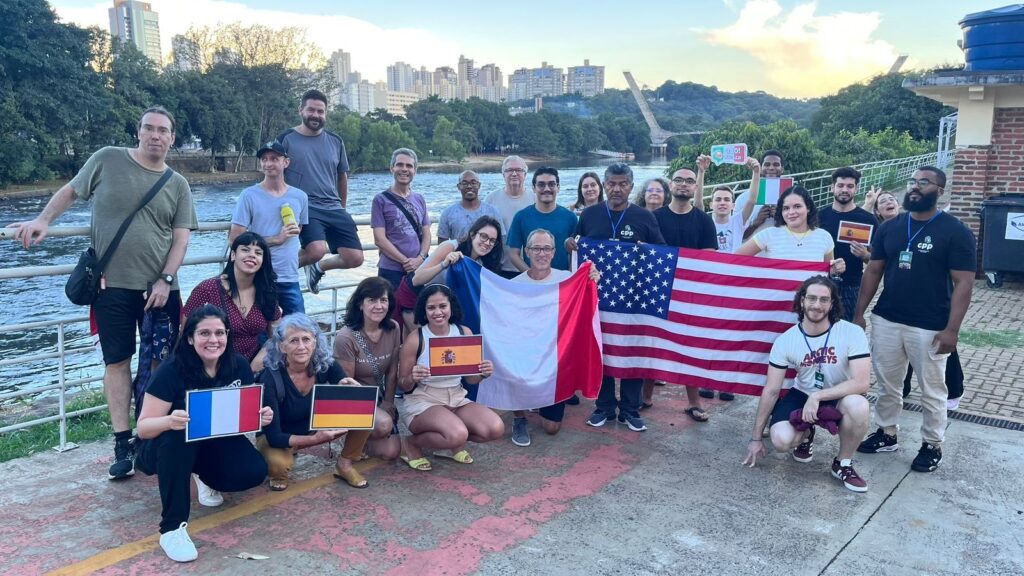 A imagem mostra um grupo de cerca de 20 pessoas reunido à beira de um rio, em um espaço ao ar livre. Algumas seguram bandeiras da França, Alemanha, Peru, Estados Unidos e Espanha, além de uma grande bandeira branca e vermelha, provavelmente da Polônia. As pessoas parecem participar de um encontro internacional ou atividade cultural. Ao fundo, há árvores, um rio com corredeiras, uma ponte e vários prédios altos da cidade. À direita, aparece parte de uma construção de tijolos, e o grupo posa sorrindo para a foto.