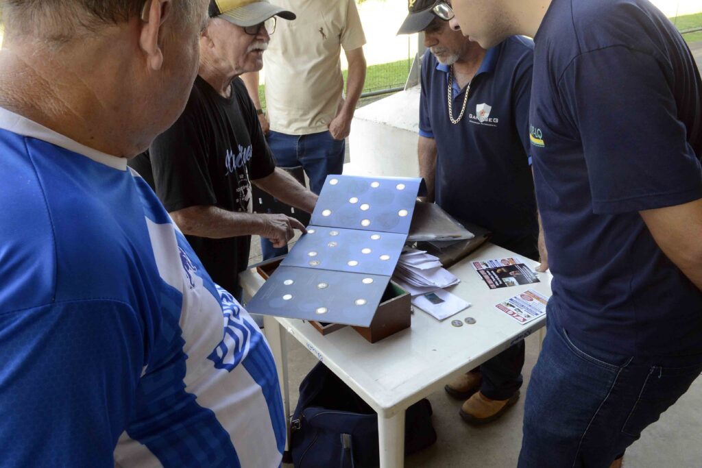 Mostra um grupo de pessoas reunidas em torno de uma mesa analisando um painel com moedas organizadas em compartimentos. A cena sugere uma atividade de coleção, troca ou avaliação de moedas durante a feira.