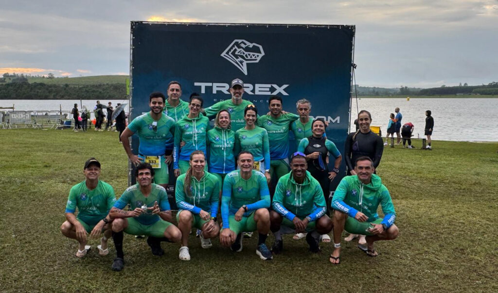 Grupo de atletas posa para uma foto em um evento de triatlo ao ar livre, em frente a um painel da competição e próximo a um lago.