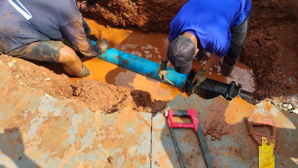 A imagem mostra dois trabalhadores realizando o reparo de uma tubulação em uma vala escavada. Eles estão dentro de uma área alagada por água barrenta, conectando um tubo azul a uma peça preta da rede.