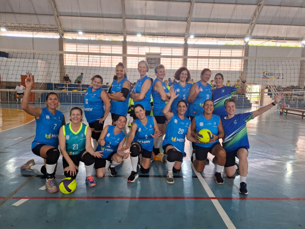 Uma equipa feminina de voleibol (categoria 45+) posa sorridente no pavilhão após garantir a classificação para a final regional. As jogadoras, vestidas com equipamentos azuis e verdes, estão distribuídas em duas filas (uma ajoelhada e outra de pé) em frente à rede de voleibol, com algumas a segurar bolas e a gesticular de forma festiva.
