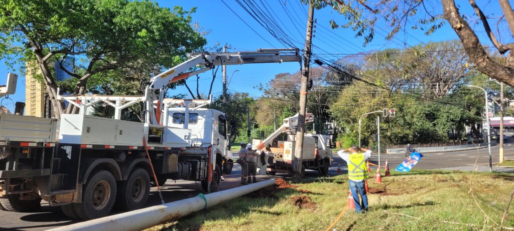 A imagem mostra uma equipe realizando uma obra de manutenção em uma via urbana arborizada. Há dois caminhões equipados com braços hidráulicos e cestos de trabalho, além de um grande tubo branco apoiado no gramado e conectado à área escavada. Os trabalhadores usam uniformes com faixas refletivas e capacetes. Cones laranjas sinalizam o serviço, enquanto árvores, postes e muitos cabos elétricos aparecem ao redor. Ao fundo, há um semáforo, uma avenida movimentada e prédios residenciais.