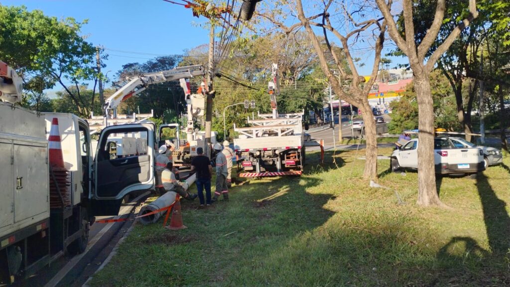 A imagem mostra uma equipe de trabalhadores realizando manutenção em uma área gramada próxima a uma avenida. Eles usam uniformes com faixas refletivas e capacetes, enquanto operam caminhões equipados com cestos e braços hidráulicos. Há cones de sinalização, ferramentas e materiais espalhados pelo local. Os trabalhadores parecem atuar junto a postes e cabos elétricos. Ao fundo, vê-se uma via movimentada, semáforos, árvores e alguns estabelecimentos comerciais.