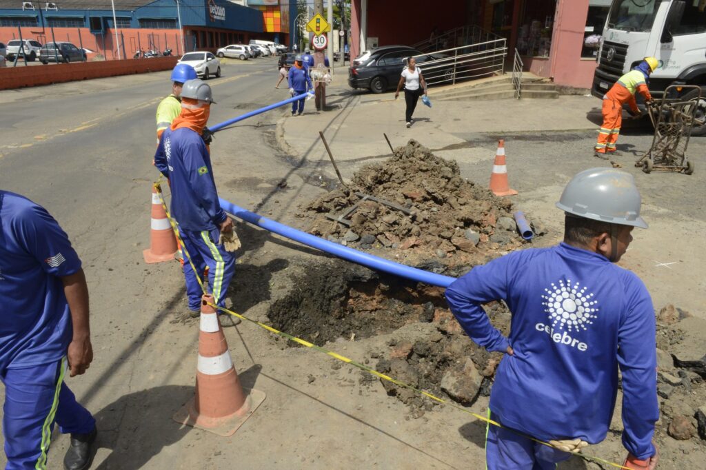 Equipes em serviço ao redor de uma escavação na via pública, posicionando tubulações e realizando intervenções na rede de água, com cones e fitas de segurança delimitando a área de trabalho.