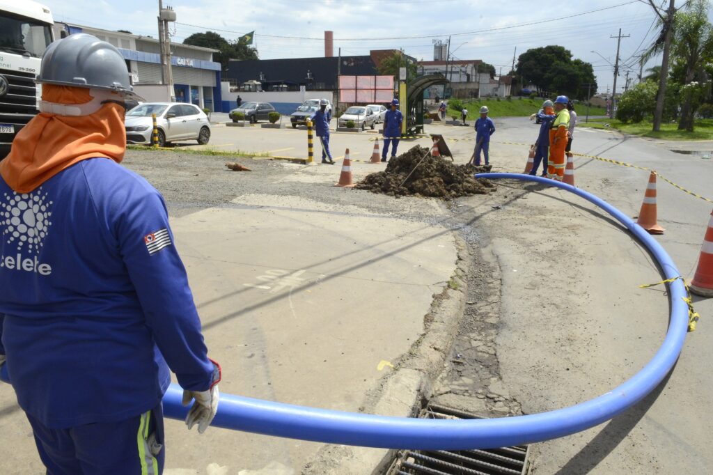 Trabalhadores atuam em uma obra de infraestrutura, transportando uma tubulação azul de grande porte para a implantação de uma nova rede de abastecimento de água.