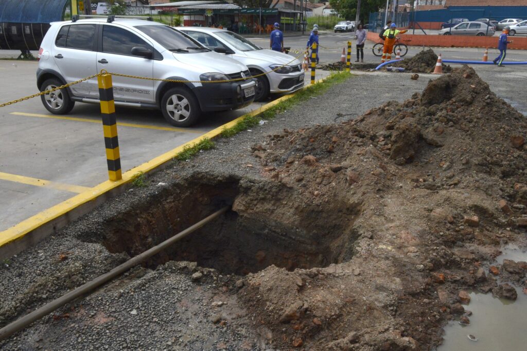 Escavação realizada em uma área urbana para instalação ou substituição de tubulações de água. O local está sinalizado e há montes de terra ao lado da vala aberta.