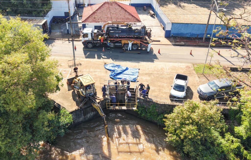 A imagem mostra uma operação de limpeza ou manutenção de drenagem urbana vista de cima. Há uma equipe de trabalhadores atuando sobre uma estrutura metálica instalada na margem de um rio ou canal com água barrenta.