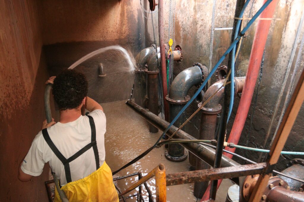 A imagem mostra um trabalhador de saneamento realizando limpeza de sistema de bombeamento ou drenagem. Ele usa equipamentos de proteção — calça impermeável amarela e camiseta branca — e segura uma mangueira que lança jato d’água sobre as paredes internas.