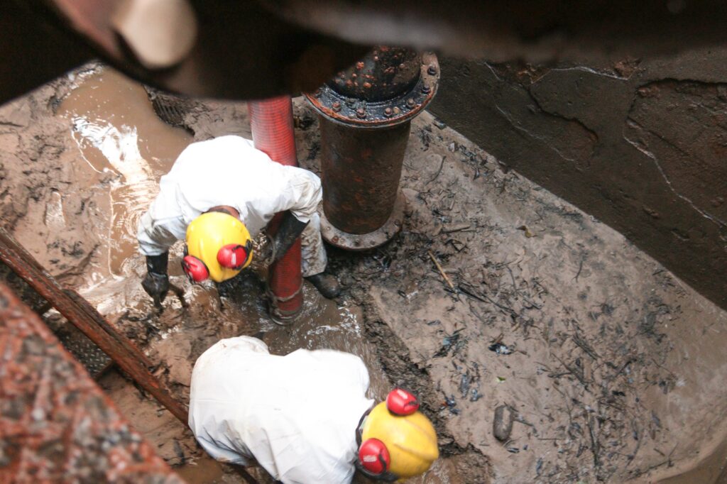 A imagem mostra dois trabalhadores de saneamento realizando limpeza de tubulação em um ambiente confinado e lamacento. Eles vestem macacões brancos impermeáveis, capacetes amarelos e protetores auriculares vermelhos, indicando o uso de equipamentos de segurança.