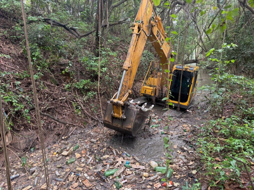 A imagem mostra uma escavadeira hidráulica trabalhando na limpeza de um canal em meio a uma área de mata. O equipamento está posicionado no leito do curso d’água, cercado por vegetação densa.
