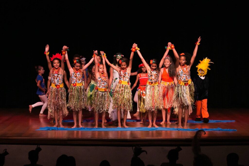 A imagem mostra um grupo de crianças e adolescentes em uma apresentação teatral sobre um palco. Os participantes usam figurinos coloridos inspirados na natureza, com saias de franjas, flores e acessórios temáticos, enquanto alguns personagens ao fundo vestem fantasias especiais.