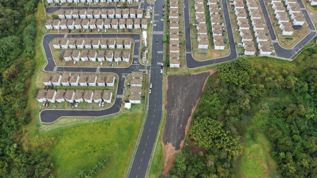 A imagem aérea mostra um conjunto habitacional planejado, cercado por áreas verdes e mata. Há dezenas de casas brancas, muito semelhantes entre si, organizadas em fileiras e quadras. As ruas são asfaltadas, bem sinalizadas e possuem faixas de pedestres, calçadas e algumas vagas de estacionamento. Também é possível ver pequenos parques ou áreas de lazer entre as residências. Uma avenida principal atravessa o empreendimento, com alguns carros circulando. À direita, há um grande terreno de solo exposto e vegetação densa; à esquerda, uma extensa área de mata.