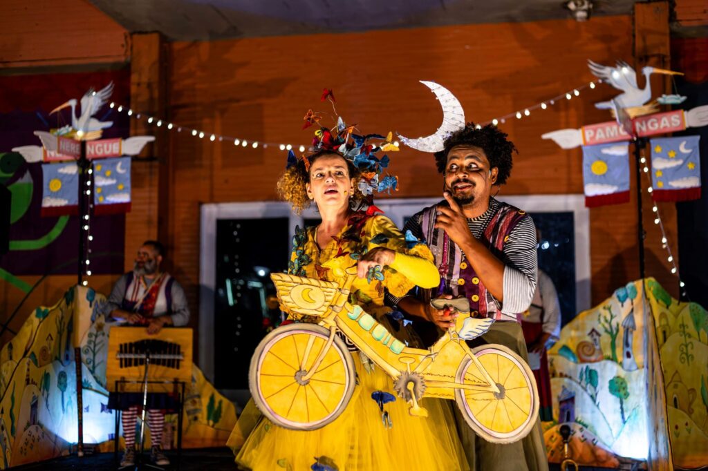 A imagem mostra uma cena teatral cheia de cor e fantasia. No centro, dois artistas estão em destaque: a mulher veste um figurino amarelo vibrante decorado com borboletas e segura uma bicicleta cenográfica amarela com asas e símbolos de lua e sol; ao lado dela, o homem usa roupas listradas e um colete colorido, com uma meia-lua presa à cabeça, sugerindo um personagem lúdico.
