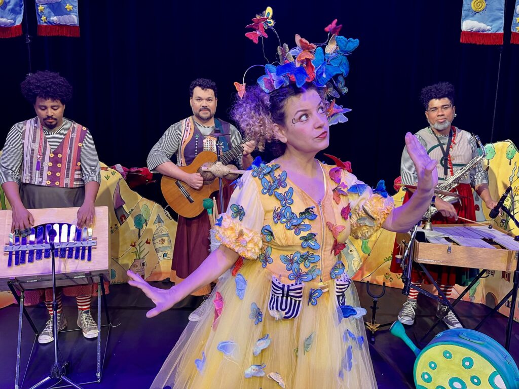 A imagem mostra uma cena teatral vibrante e cheia de fantasia. No centro, uma mulher usa um vestido amarelo decorado com dezenas de borboletas coloridas e um adorno de cabeça exuberante com flores e borboletas, transmitindo leveza e magia. Atrás dela, três músicos tocam instrumentos — um xilofone artesanal, um violão e um saxofone — todos vestidos com figurinos listrados e coloridos, que reforçam o clima lúdico da apresentação.