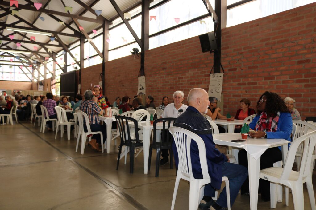 Pessoas idosas participam de um encontro comunitário em um salão amplo, sentadas em mesas e interagindo durante uma programação social e recreativa.
