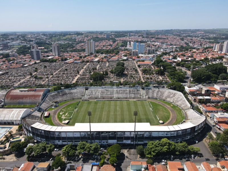 Vista aérea de um estádio de futebol e um cemitério adjacente numa cidade ensolarada.
A imagem mostra uma vista aérea de um estádio de futebol com arquibancadas e um campo verde bem cuidado, com linhas brancas visíveis. O estádio tem uma estrutura circular com áreas de assentos a céu aberto e cobertas, e holofotes nas extremidades. Ao lado do estádio, estende-se um vasto cemitério, repleto de fileiras e fileiras de túmulos e monumentos funerários. O cemitério é um grande espaço de terra coberto por inúmeras lápides e cruzes, com algumas árvores esparsas.
O ambiente em redor do estádio e cemitério é uma paisagem urbana, com edifícios residenciais de telhados laranja e alguns edifícios mais altos de múltiplos andares visíveis ao longe. Árvores verdes pontuam a paisagem urbana e circundam partes do estádio. O céu está limpo e de um azul claro, sugerindo um dia ensolarado. A iluminação é brilhante e uniforme, lançando sombras definidas, o que indica que o sol está alto no céu. A atmosfera geral é serena e um pouco melancólica, com o contraste entre o local de desporto e entretenimento e o local de descanso final. A composição da imagem é dominada pelo estádio e pelo cemitério, que ocupam a maior parte do quadro, com a cidade a estender-se para o fundo sob o céu claro.