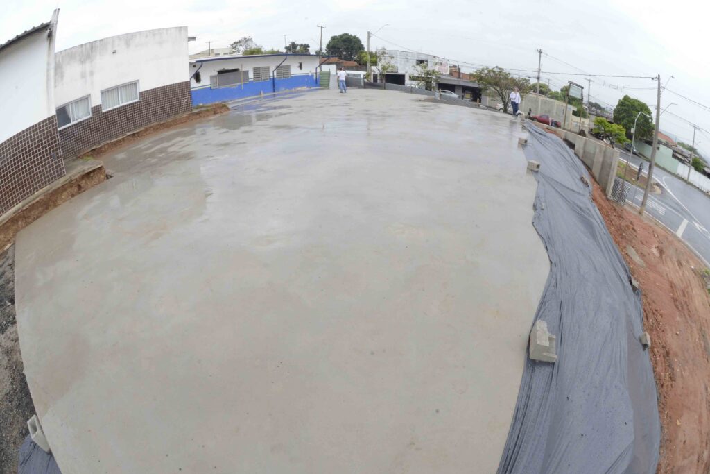 A imagem mostra uma área externa com a marcação de uma construção onde o concreto fresco foi recém-aplicado. A imagem captura um espaço aberto em um dia nublado, mostrando uma grande área de concreto recém-derramado que ainda parece úmido, com algumas ondulações sugerindo que está em processo de secagem. Ao fundo, à esquerda, vislumbra-se um edifício com uma fachada branca e detalhes em marrom quadriculado, além de janelas com cortinas brancas. Em frente a este edifício, há uma área com azul vibrante na parte inferior da parede. Mais ao centro e à direita, outras construções aparecem, algumas com aparência mais antiga e outras em diferentes estágios de reforma ou construção. Uma rua com faixas de pedestres é visível à direita, com postes de iluminação e fiação elétrica cruzando o céu de tom claro e uniforme, indicando um dia sem sol. Algumas pessoas vestidas com roupas casuais transitam pela cena, uma delas caminhando em direção à edificação azul, enquanto outra, uma mulher, caminha mais ao fundo na área da rua. Parapeitos de concreto e terra avermelhada compõem a lateral direita da imagem, com uma manta plástica cinza cobrindo uma área de aterro, possivelmente para proteção contra erosão. A atmosfera geral da imagem sugere um ambiente urbano em processo de desenvolvimento ou revitalização, com um ar de trabalho em andamento. A iluminação difusa do céu nublado contribui para uma sensação de normalidade e continuidade.