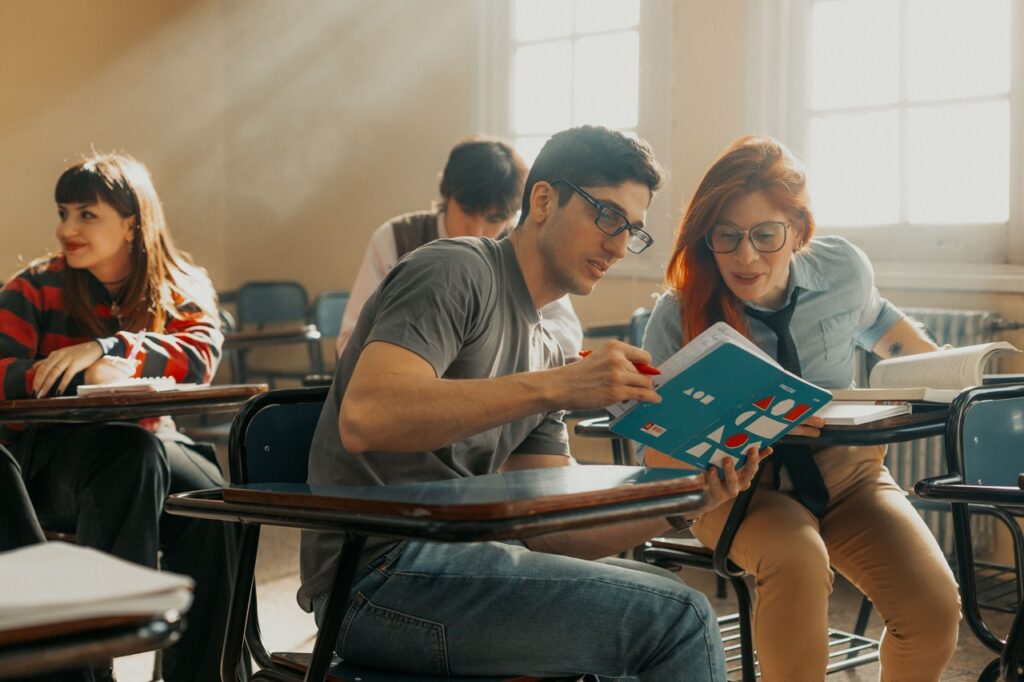 Sala de aula com estudantes sentados em carteiras, consultando materiais de estudo e interagindo durante uma atividade acadêmica em ambiente escolar.