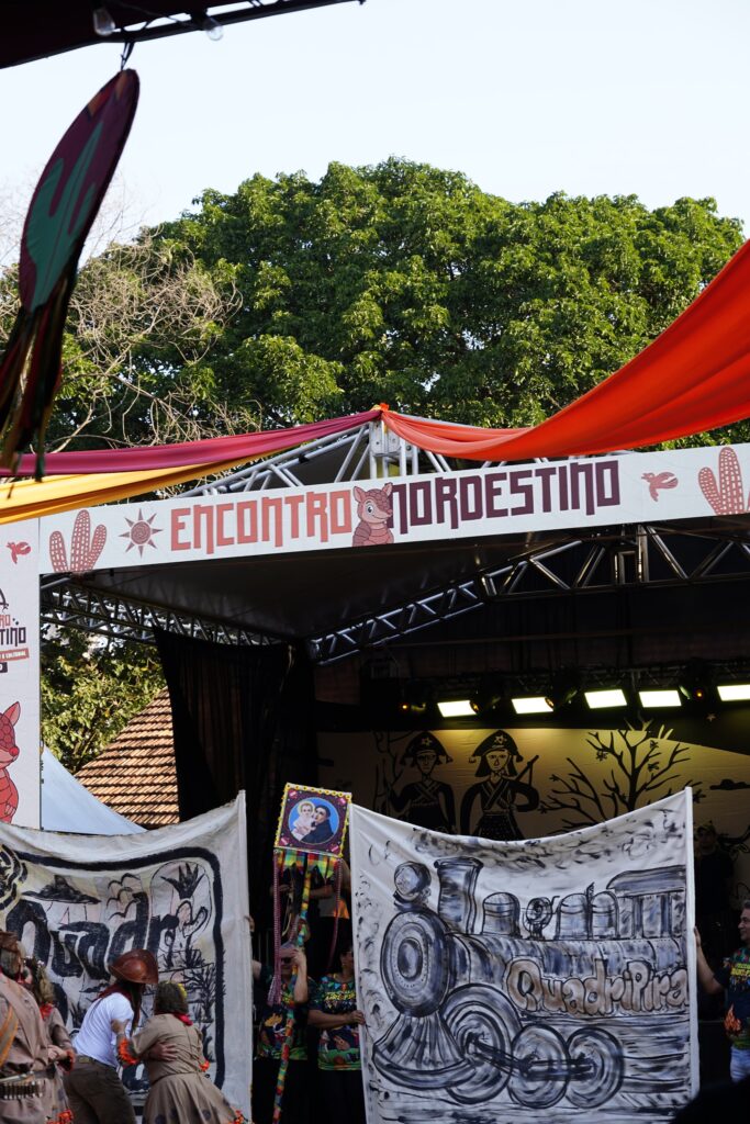A imagem mostra um evento cultural chamado Encontro Nordestino, realizado em um palco decorado com elementos típicos da cultura do Nordeste brasileiro — como cactos, sol e um tatu estilizado. Há tecidos coloridos (vermelho e laranja) formando uma cobertura sobre o palco, e ao fundo aparecem árvores e parte de um telhado.