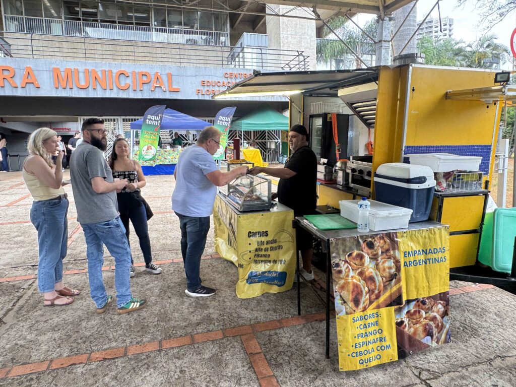 A imagem mostra uma barraca de comida em uma área externa, diante de um prédio identificado como Centro Educacional Floripa. Há várias pessoas aguardando ou fazendo compras. À direita, um vendedor de roupa preta atende um homem de camisa azul diante de um balcão com alimentos. A barraca é amarela e tem cartazes anunciando empanadas argentinas, com sabores como carne, frango, espinafre, cebola e queijo. Também aparecem caixas térmicas, recipientes plásticos e uma vitrine com produtos. Ao fundo, há tendas coloridas, cartazes de cocadas e uma fachada com grades e janelas.