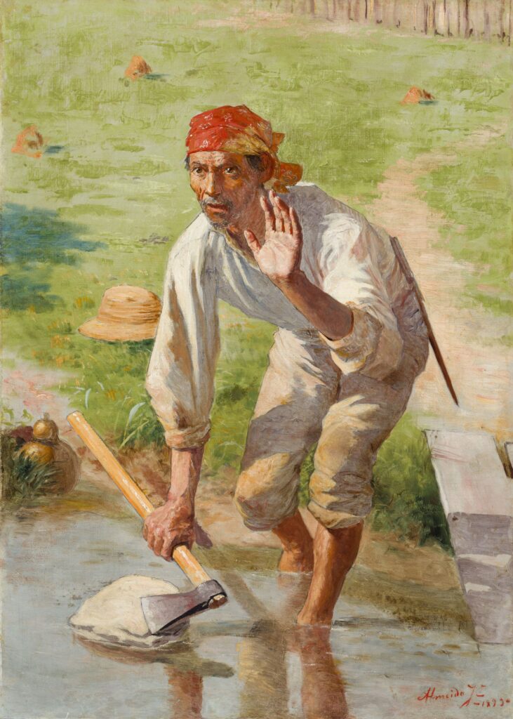 imagem mostra um trabalhador rural ajoelhado em um campo alagado, usando roupas simples e um lenço vermelho na cabeça. Ele segura uma enxada e parece trabalhar em uma plantação, enquanto a paisagem verde e o reflexo na água compõem a cena.