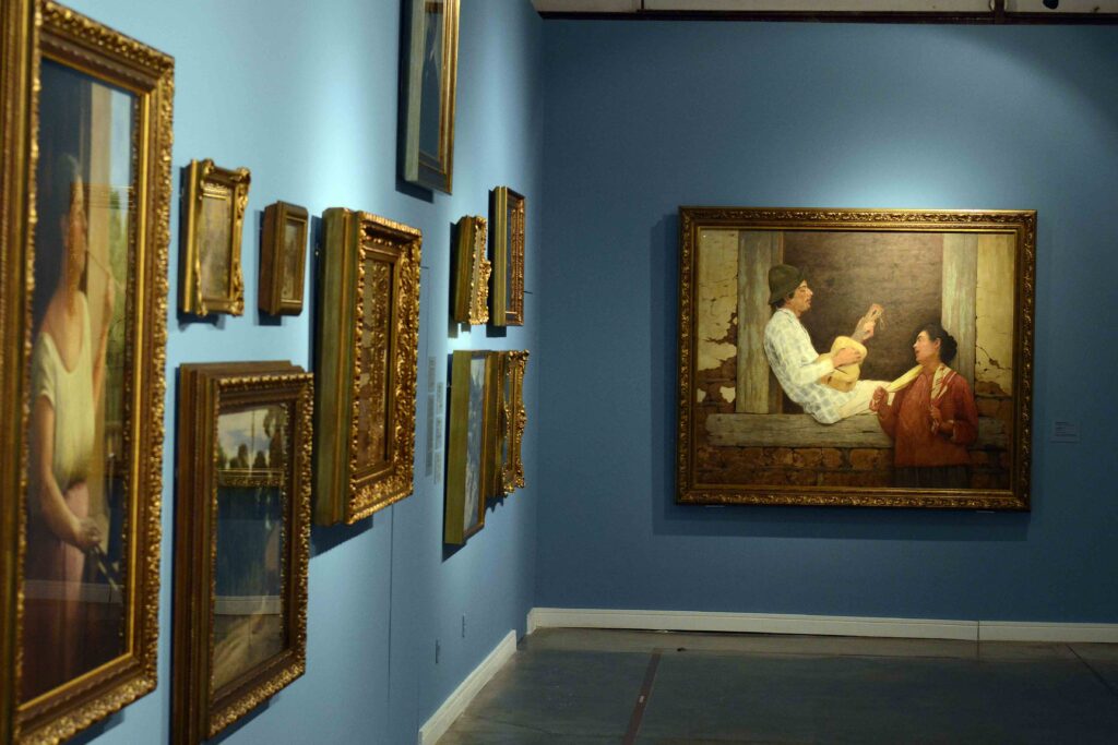 Sala de exposição com diversas obras distribuídas pelas paredes azuis, incluindo uma pintura maior ao fundo retratando duas pessoas em uma cena cotidiana.