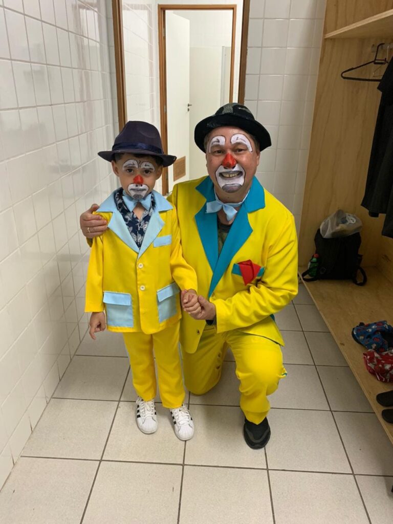 Um palhaço adulto e uma criança vestem trajes de palhaço semelhantes e pintam o rosto, compartilhando um momento de união e alegria antes de uma apresentação. A imagem mostra um palhaço adulto ajoelhado e um menino em pé, ambos vestidos com roupas de palhaço vibrantes e combinando. O palhaço adulto usa um terno amarelo com detalhes em azul, um chapéu preto e uma gravata borboleta azul. Sua maquiagem de palhaço é clássica, com o rosto pintado de branco, um nariz vermelho e detalhes em preto e vermelho ao redor dos olhos e boca, que está aberta em um sorriso largo. O menino, que parece ser filho do adulto, veste um terno amarelo semelhante com detalhes em azul claro, um chapéu azul escuro e uma gravata borboleta azul estilizada. Sua maquiagem de palhaço também inclui a base branca, nariz vermelho e detalhes expressivos em preto e vermelho. Eles estão de mãos dadas, com o braço do adulto em volta dos ombros do menino, transmitindo um senso de orgulho e afeto. O cenário parece ser um vestiário ou corredor dos fundos. As paredes são revestidas com azulejos brancos. À direita, há um armário de madeira com cabides e roupas penduradas, incluindo um vestido preto. Abaixo, há alguns objetos no chão, incluindo uma mochila escura e sacolas. A iluminação é clara e suave, iluminando os sujeitos e o ambiente. A atmosfera da imagem é calorosa e alegre. A combinação das roupas coloridas, a maquiagem de palhaço e a interação afetuosa entre o adulto e a criança criam um sentimento de diversão, antecipação e carinho familiar. Há uma sensação de que eles estão prestes a realizar algo especial juntos e que compartilham um vínculo forte através de sua arte. O humor é otimista e encantador, sugerindo a alegria e a magia que os palhaços trazem.