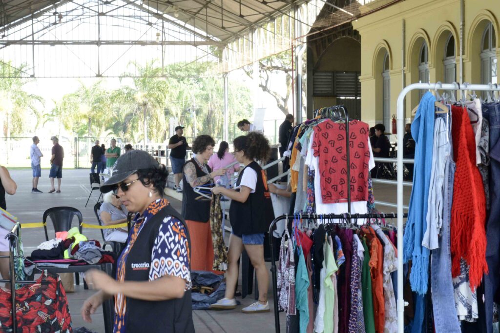 Mostra uma feira de troca e reutilização em um espaço coberto. Há várias araras com roupas coloridas, incluindo camisetas, calças e cachecóis. Diversas pessoas circulam pelo local, observando e organizando os itens expostos.