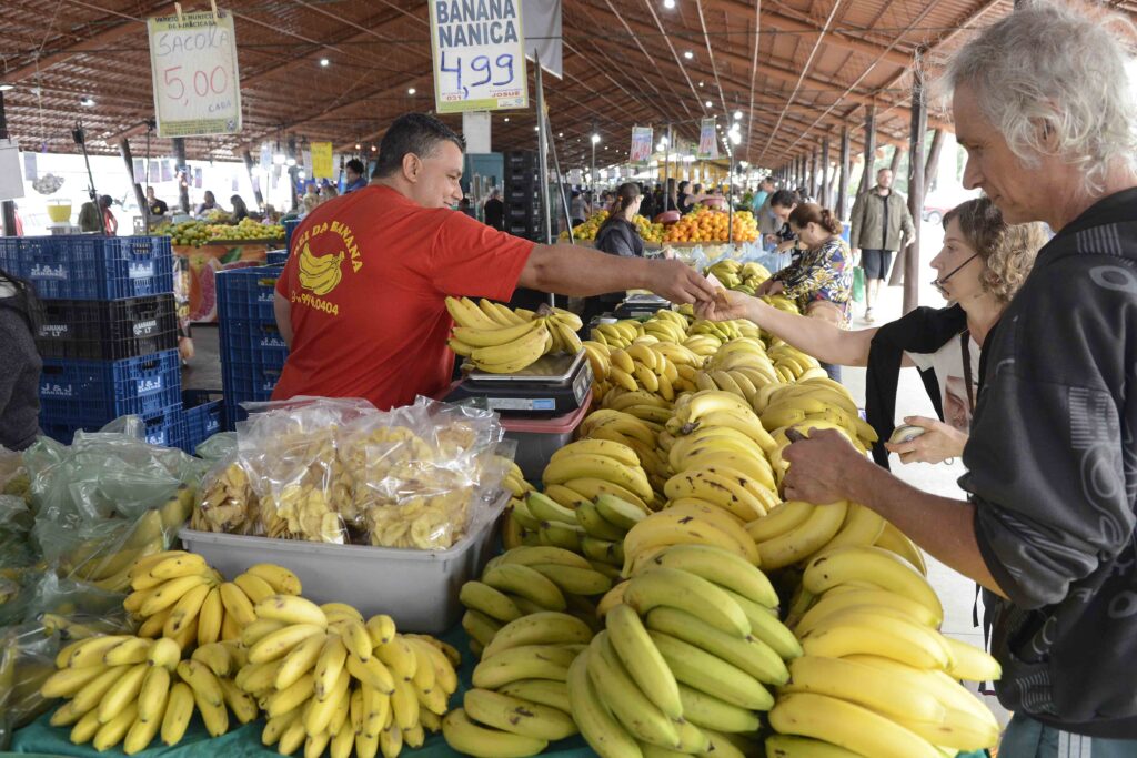 Feirante atende clientes em uma banca de bananas dentro de um mercado municipal, com grande variedade de frutas expostas e placas de preços visíveis ao fundo.