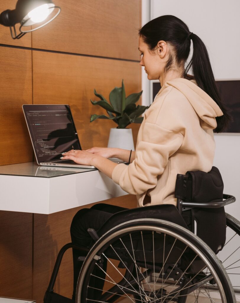 A imagem mostra uma pessoa utilizando um notebook em uma mesa de trabalho, com a tela exibindo linhas de código. A pessoa está sentada em uma cadeira de rodas, em um ambiente interno organizado e bem iluminado.