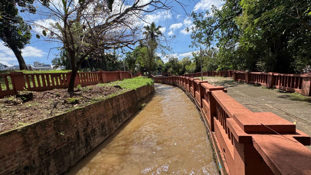 A imagem mostra um canal de água com correnteza moderada atravessando uma área arborizada. O canal é ladeado por muretas e guarda-corpos vermelhos, ao lado de uma calçada pavimentada.