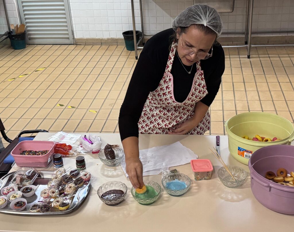 Participante de uma aula prática de confeitaria prepara mini donuts em uma cozinha, mergulhando os produtos em coberturas coloridas e utilizando confeitos para decoração.