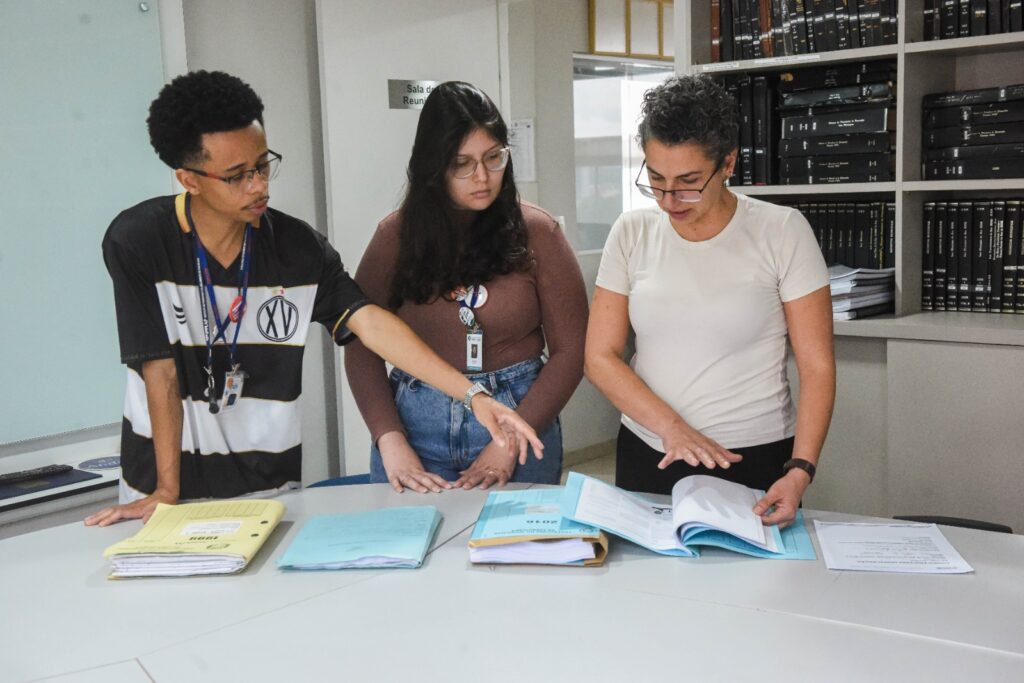 A imagem mostra três pessoas reunidas em um ambiente de trabalho ou estudo, analisando documentos sobre uma mesa. Elas parecem concentradas, apontando e discutindo o conteúdo das pastas e folhas abertas.
