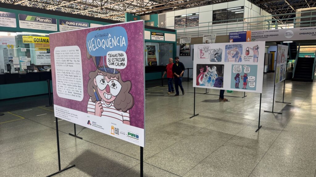 Exposição de tirinhas e cartuns em painéis montados dentro de uma rodoviária, com visitantes circulando pelo espaço.