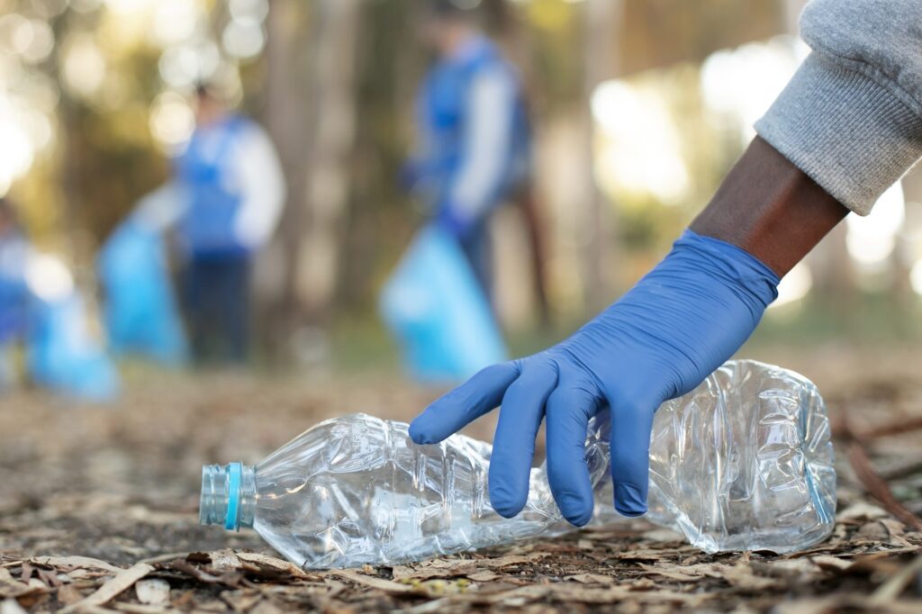 A imagem mostra uma pessoa usando uma luva azul descartável enquanto recolhe uma garrafa plástica vazia do chão, em uma área arborizada. Ao fundo, outras pessoas, também usando luvas e roupas em tons de azul, participam da limpeza, mas aparecem desfocadas. O solo está coberto por folhas secas, e a cena destaca uma ação de coleta de resíduos e cuidado com o meio ambiente.