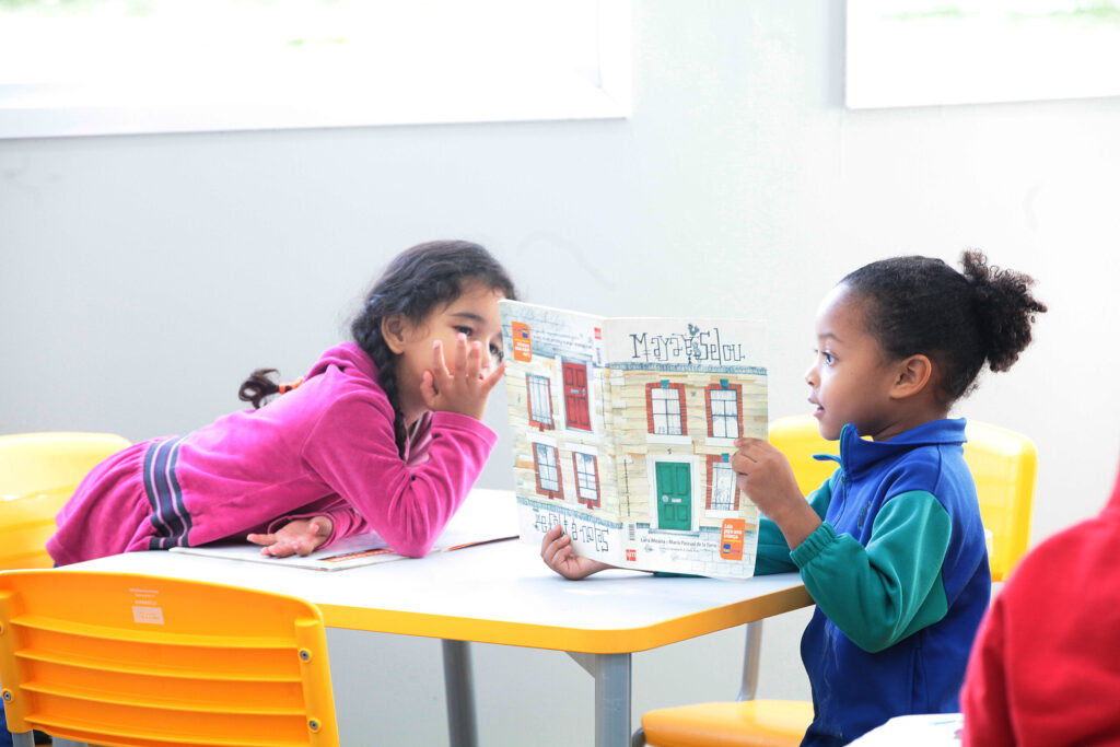 Duas crianças pequenas estão sentadas a uma mesa amarela numa sala de aula, compartilhando e lendo juntas um livro infantil ilustrado.