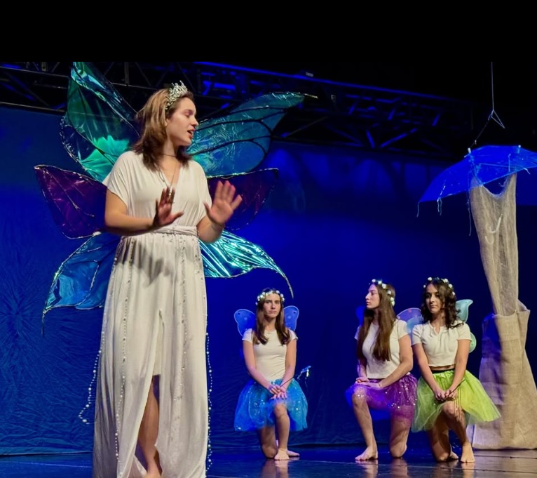 A imagem mostra uma cena teatral em um palco iluminado por luz azul. Em primeiro plano, uma pessoa veste uma roupa branca longa e grandes asas coloridas de fada.