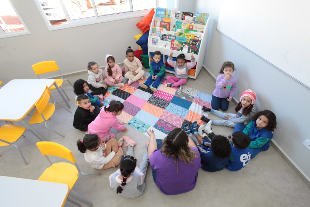 Um grupo de crianças da educação infantil e a sua professora estão dispostos em roda sobre um tapete colorido no chão da sala de aula, ao lado de uma estante de livros.