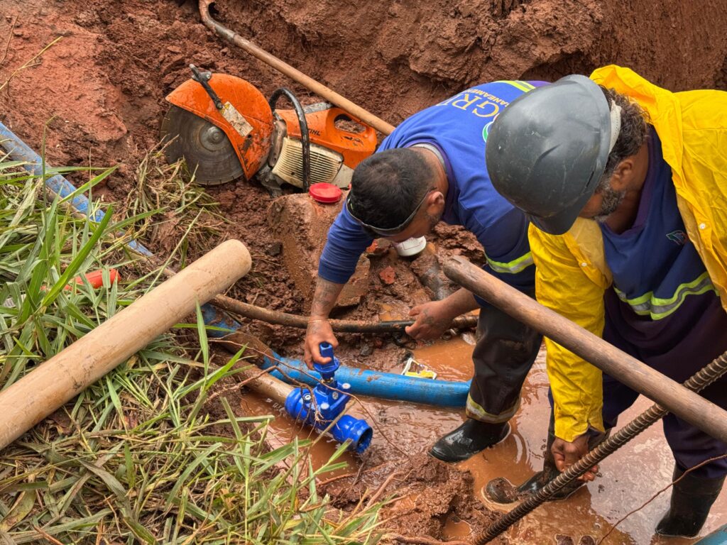 A imagem mostra uma equipe trabalhando em uma área de terra enlameada, aparentemente para reparar uma tubulação de água. Duas pessoas usam uniformes de alta visibilidade amarelos e azuis; uma delas segura uma ferramenta, enquanto a outra se inclina sobre a escavação. Ao centro, há uma motosserra laranja apoiada no solo, além de um cabo ou mangueira de madeira e tubos azuis expostos. A água barrenta se acumula na vala, cercada por grama, raízes e terra revolvida.