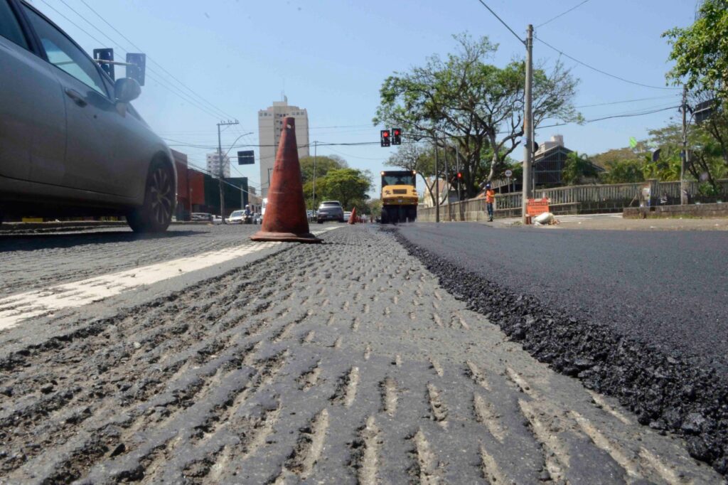 Avenida ampla com asfalto novo, cercada por áreas verdes, muros e estabelecimentos comerciais às margens da via.