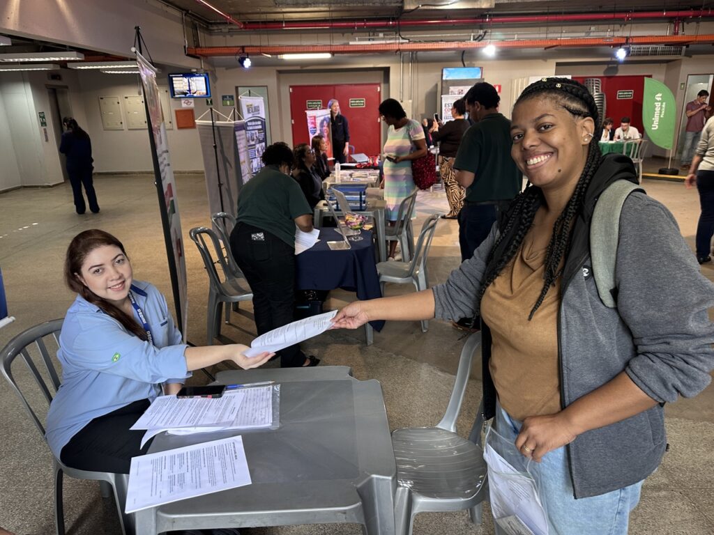 Candidata entrega documentos em um estande de atendimento durante uma feira de empregabilidade, enquanto outras pessoas circulam pelo local em busca de oportunidades de trabalho.