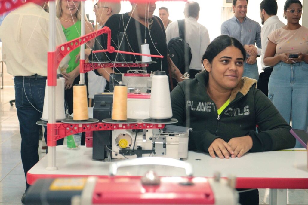 A imagem mostra uma mulher sentada diante de uma máquina de costura industrial, em um ambiente movimentado. A máquina tem estrutura branca e vermelha, com vários carretéis de linha — brancos e alaranjados — posicionados nos suportes. A mulher usa um casaco escuro com capuz e observa algo à sua frente. Ao fundo, várias pessoas conversam e acompanham a atividade, sugerindo uma feira, oficina ou evento de costura. Há outras máquinas semelhantes parcialmente visíveis em primeiro plano.