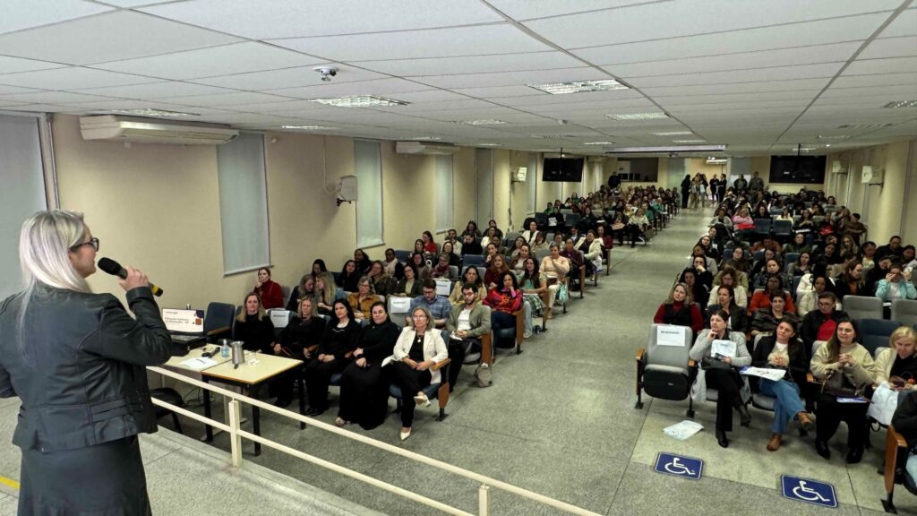 A imagem mostra uma mulher falando ao microfone diante de um auditório cheio. Ela está posicionada à esquerda, de frente para o público, e parece conduzir uma palestra ou apresentação.
