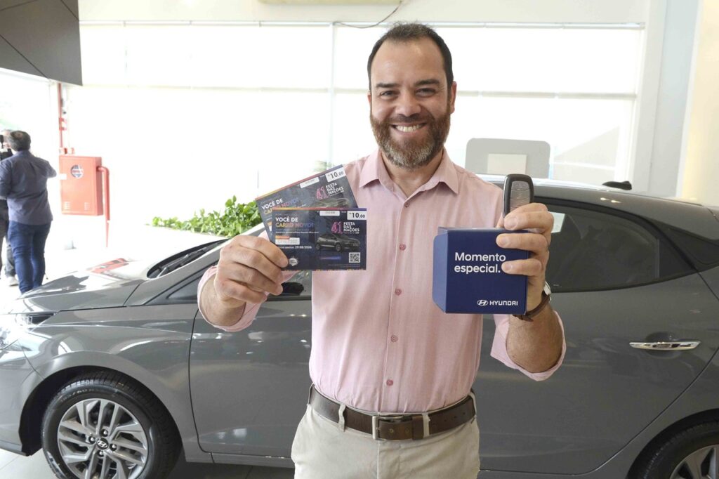 A imagem mostra um homem sorridente dentro de uma concessionária Hyundai, ao lado de um carro cinza. Ele segura uma caixa azul com a inscrição “Momento especial” e uma chave de carro, além de dois bilhetes promocionais que dizem “Você de carro novo!” e “41ª Festa das Nações”. O cenário indica um momento de celebração pela aquisição de um veículo novo, possivelmente vinculado a uma ação promocional ou sorteio.