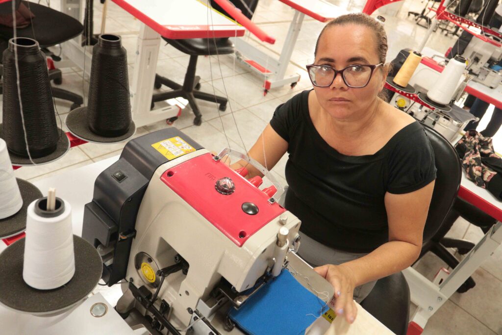 A imagem mostra duas pessoas sentadas lado a lado em uma mesa, participando de uma atividade com máquinas de costura ou bordado. A pessoa à esquerda usa óculos escuros e uma camiseta preta com estampa floral; a pessoa à direita também usa óculos e uma camiseta preta. Sobre a mesa há várias máquinas vermelhas e brancas, carretéis de linha, tecidos, papéis, ferramentas e outros materiais de artesanato. A cena parece fazer parte de uma oficina ou curso de costura.