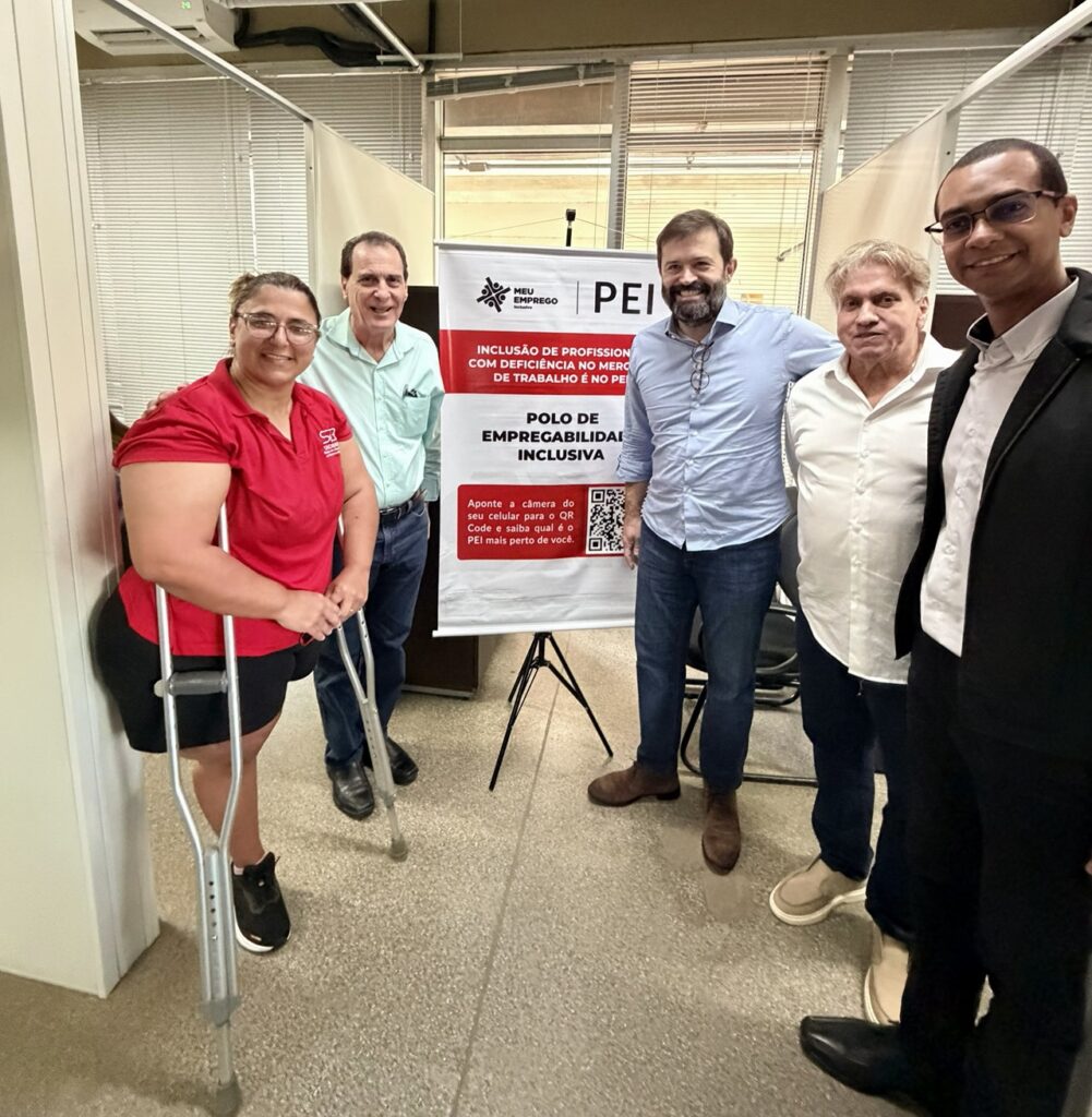 Grupo de pessoas posa para uma foto em um ambiente corporativo, ao lado de um banner sobre inclusão de profissionais com deficiência no mercado de trabalho.