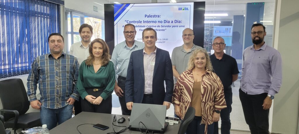 Grupo de participantes posa para uma foto em uma sala de reuniões durante uma capacitação. Ao fundo, uma tela exibe uma palestra sobre controle interno e fiscalização eficiente.