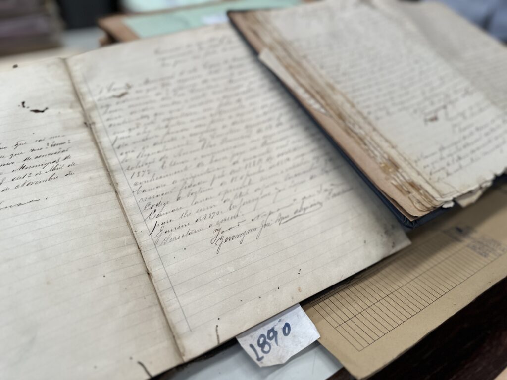 A imagem mostra um conjunto de documentos antigos dispostos sobre uma mesa. No centro, há um caderno ou livro aberto com páginas amareladas e texto escrito à mão em tinta escura, com caligrafia cursiva elegante. Um pequeno papel com o número “1890” indica o ano do registro, sugerindo tratar-se de um documento histórico.