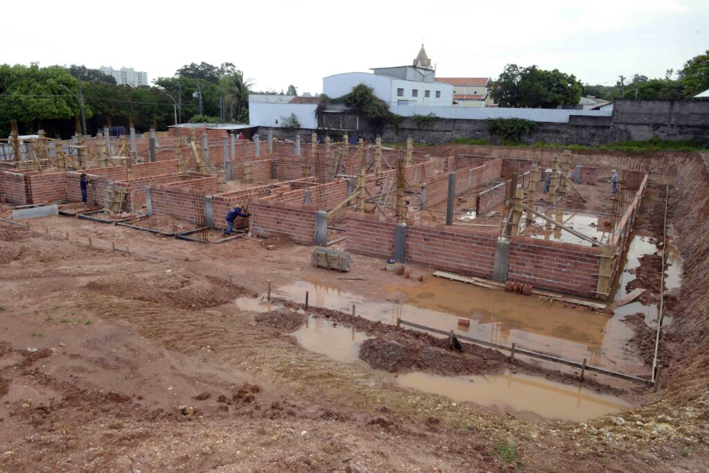 vista geral de uma grande edificação em fase de construção, com paredes de alvenaria, pilares estruturais e trabalhadores atuando no canteiro de obras.